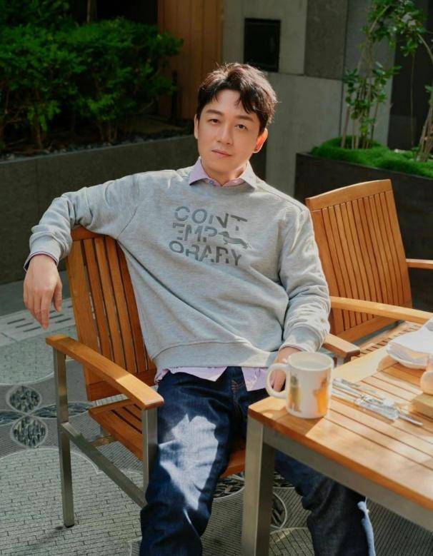 照片中潘粤明倚靠在一面绿色背景墙上,身着一件长款风衣,搭配一条宽松